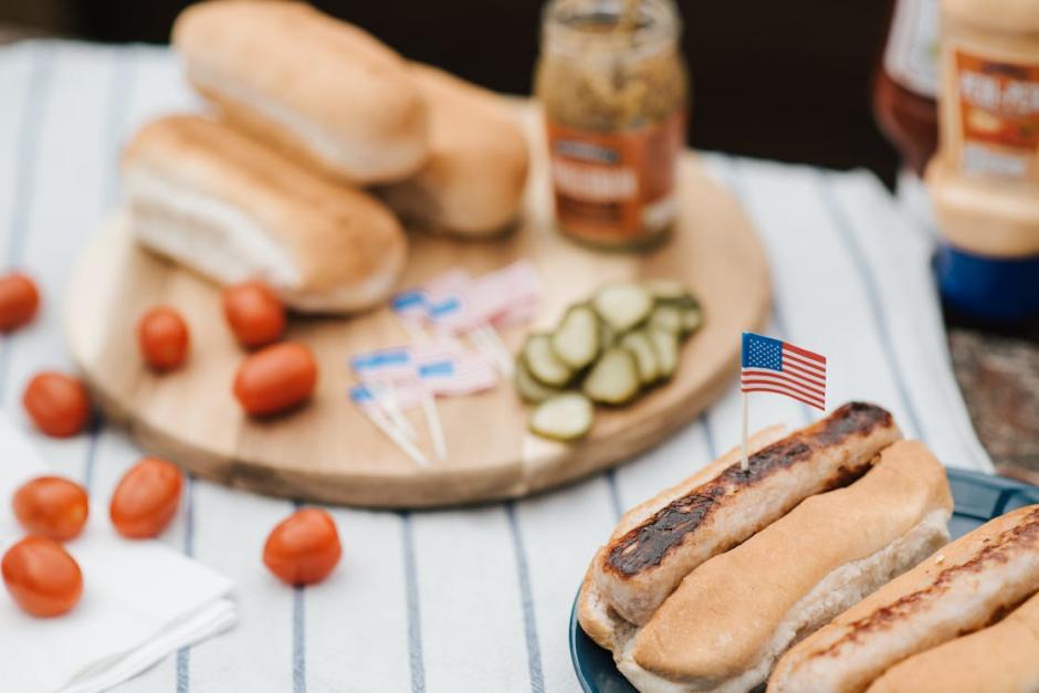Hot dogs in buns with American flag toothpicks on a blue plate, surrounded by pickles, buns, cherry tomatoes, and condiments on a picnic table