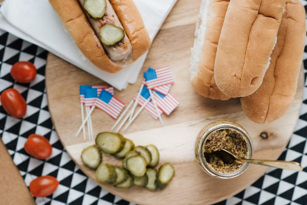 Overhead view of a pickle-topped hot dog, extra buns, and mustard on a round wooden board with small American flags and cherry tomatoes