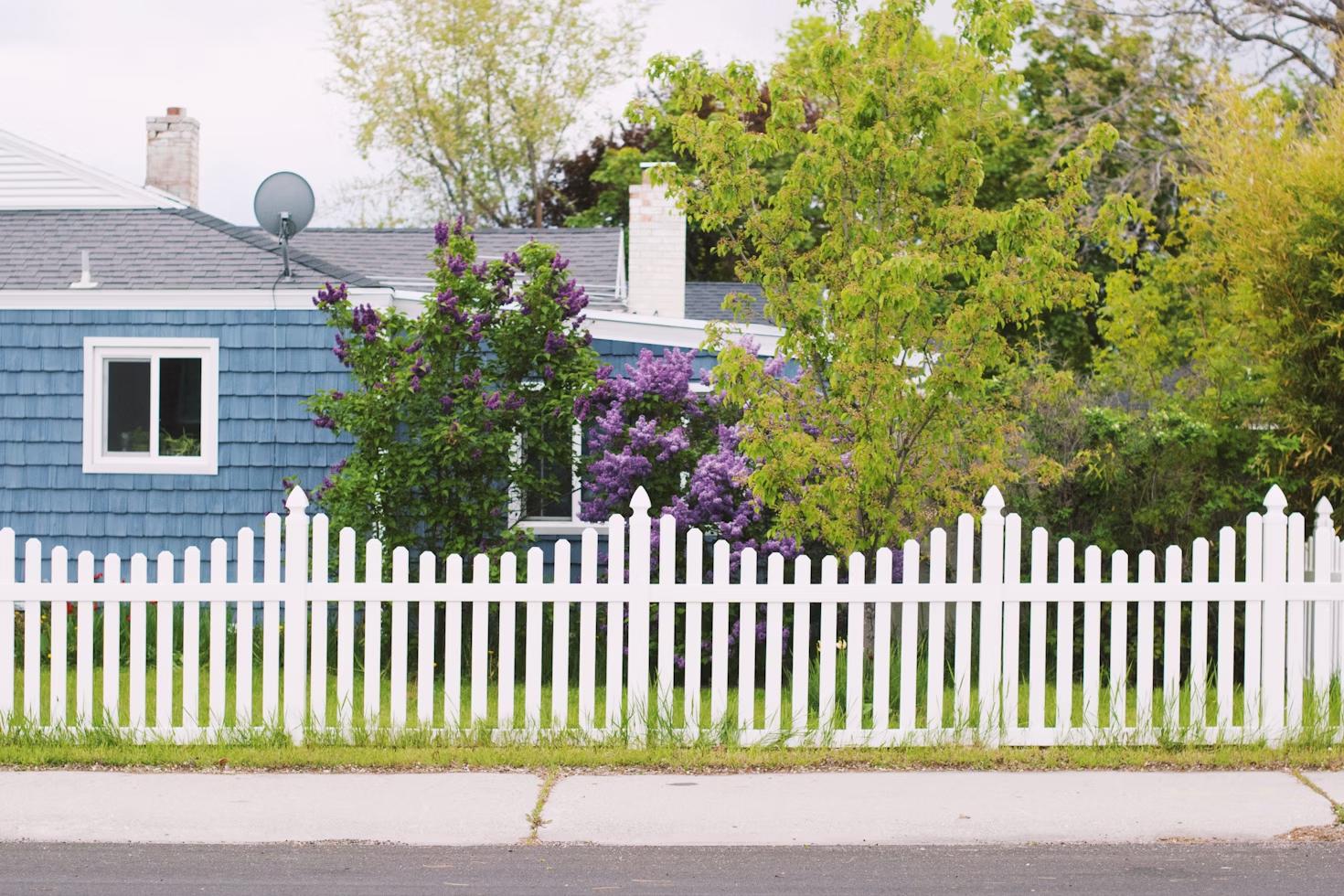 A neat white picket fence stands in front of a charming blue house with a shingle exterior.
