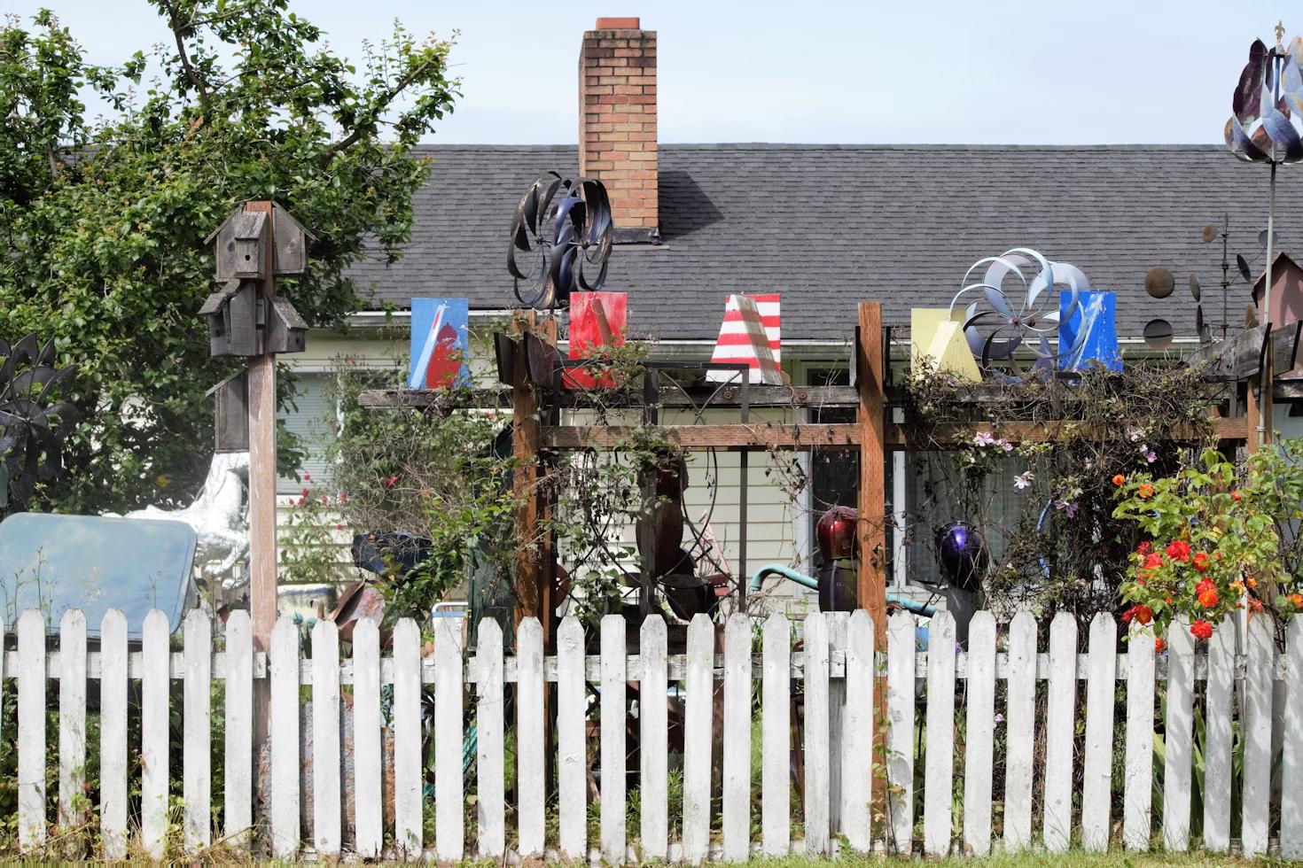 Eclectic front yard garden with a white picket fence, wooden pergola, colorful metal art, birdhouses, and blooming flowers.