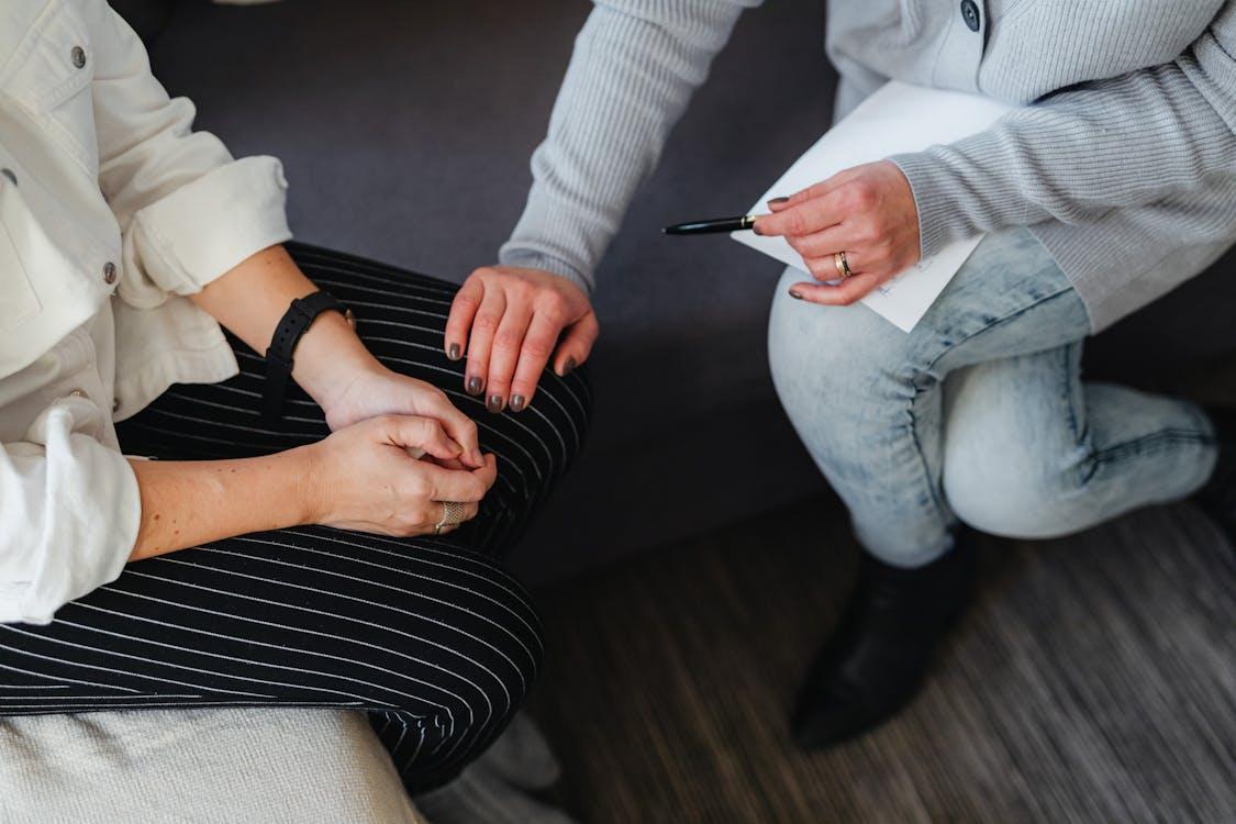 The image shows a person comforting another by gently placing a hand on their knee during what appears to be a supportive conversation or counseling session.